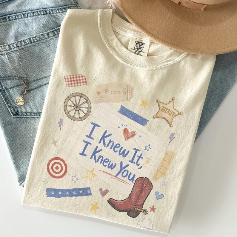 May include: A cream-colored short-sleeve t-shirt featuring a graphic print of a lined notebook page with the text "I Knew It, I Knew You" in blue. The design includes a red cowboy boot, a sheriff star, a wagon wheel, and various small icons.