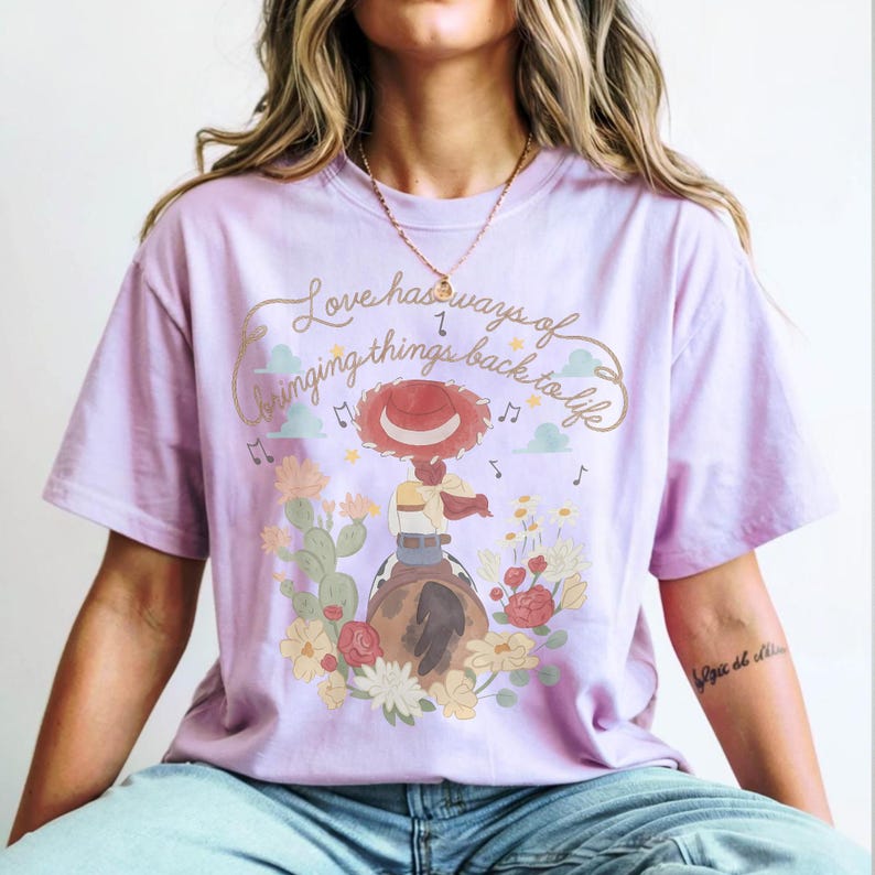 May include: A light purple short-sleeve t-shirt featuring a graphic of a cowgirl on a horse surrounded by cacti and flowers. The text reads, Love has ways of bringing things back to life, accompanied by musical notes and small clouds in the design.