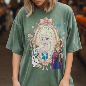 May include: Sage green t-shirt featuring a framed portrait of Elsa with Anna and Olaf. The portrait is surrounded by pink bows, flowers, and snowflakes. The shirt has a vintage, washed look.