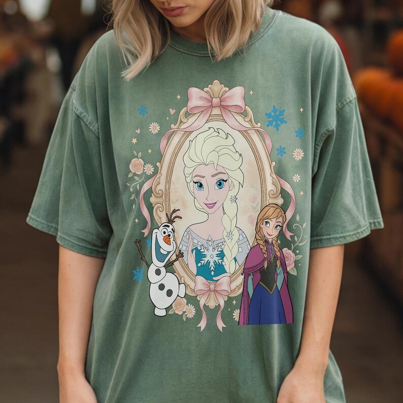 May include: Sage green t-shirt featuring a framed portrait of Elsa with Anna and Olaf. The portrait is surrounded by pink bows, flowers, and snowflakes. The shirt has a vintage, washed look.