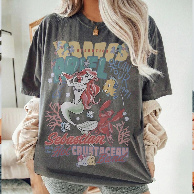 May include: Dark gray t-shirt featuring a graphic of Ariel the mermaid, Flounder, and Sebastian from The Little Mermaid. The shirt has the text "Mermaid Atlantica Ocean Tour 1991" and "Sebastian with the Hot Crustacean Band".