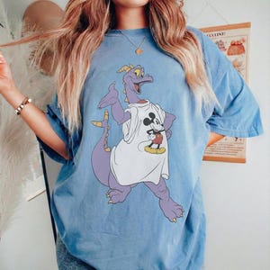Peut inclure: T-shirt bleu clair avec un motif de dragon violet portant un t-shirt blanc avec Mickey Mouse. Le dragon a des d&eacute;tails jaunes et est dans une pose dynamique. Le t-shirt a des manches courtes et une coupe ample.