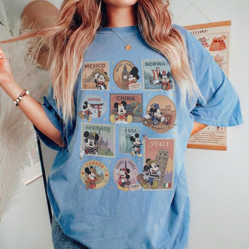 May include: Light blue t-shirt featuring Mickey Mouse postage stamp designs from various countries, including Mexico, Japan, Germany, and Italy. The stamps showcase Mickey in different travel-themed poses. The shirt has short sleeves.