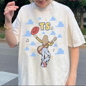 May include: A cream-colored t-shirt featuring a graphic illustration of a person with long hair wearing a yellow plaid shirt and cow-print bell-bottom pants. The person holds a guitar and a red hat, with the text TS, blue clouds, and musical notes.