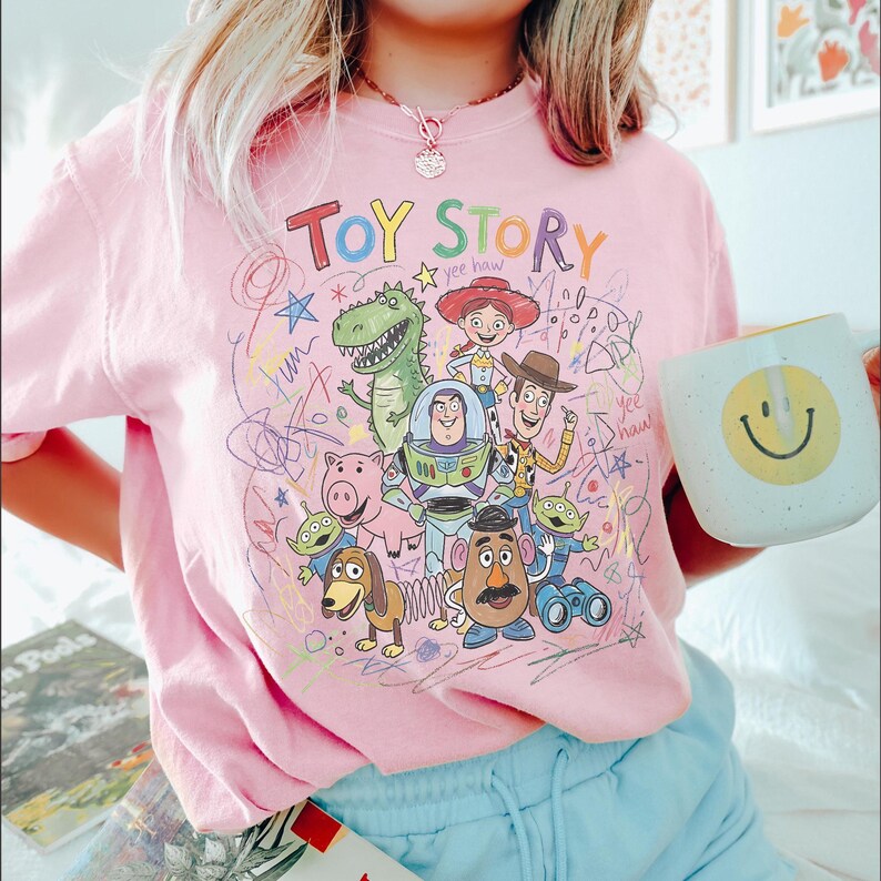 May include: A light pink t-shirt featuring a colourful graphic print of Toy Story characters including Woody, Buzz Lightyear, Jessie, Rex, Hamm, Slinky Dog, Mr. Potato Head, and Aliens, surrounded by scribbled drawings and the text Toy Story.