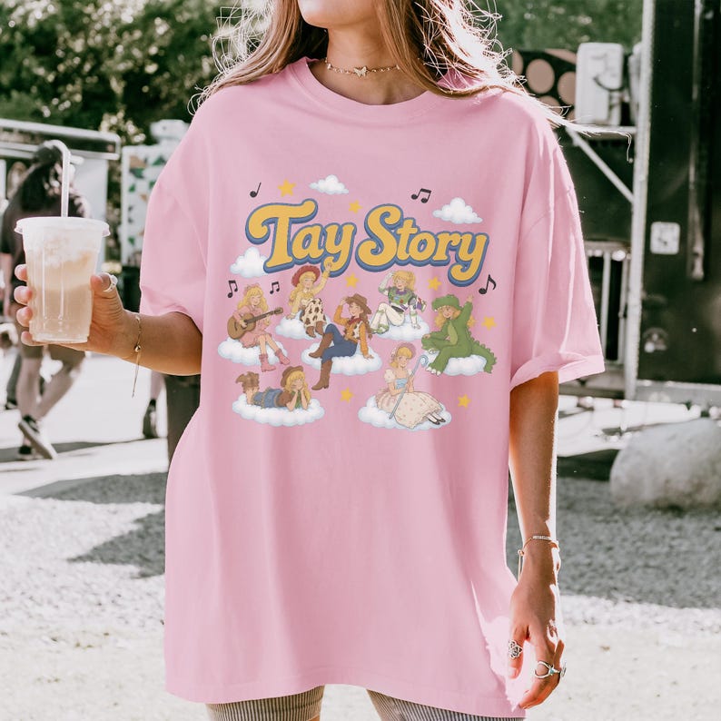 May include: A light pink t-shirt featuring the text Tay Story in yellow with a blue outline. The graphic includes multiple illustrated figures dressed in various costumes, each sitting on a white cloud, surrounded by small stars and musical notes.