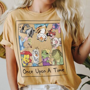 May include: Mustard yellow t-shirt featuring a square graphic with cartoon characters from various animated films. The graphic includes a raccoon, tiger, pig, and other characters. The text "Once Upon A Time" is printed below the graphic.