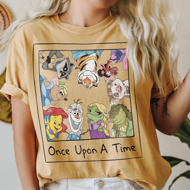 May include: Mustard yellow t-shirt featuring a square graphic with cartoon characters from various animated films. The graphic includes a raccoon, tiger, pig, and other characters. The text "Once Upon A Time" is printed below the graphic.