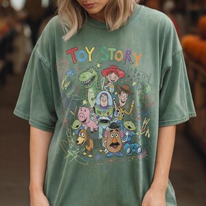 May include: A sage green short-sleeve t-shirt featuring a colourful graphic print of Toy Story characters including Woody, Buzz Lightyear, Jessie, Rex, Hamm, Slinky Dog, Mr. Potato Head, and Aliens. The text TOY STORY appears above the characters.