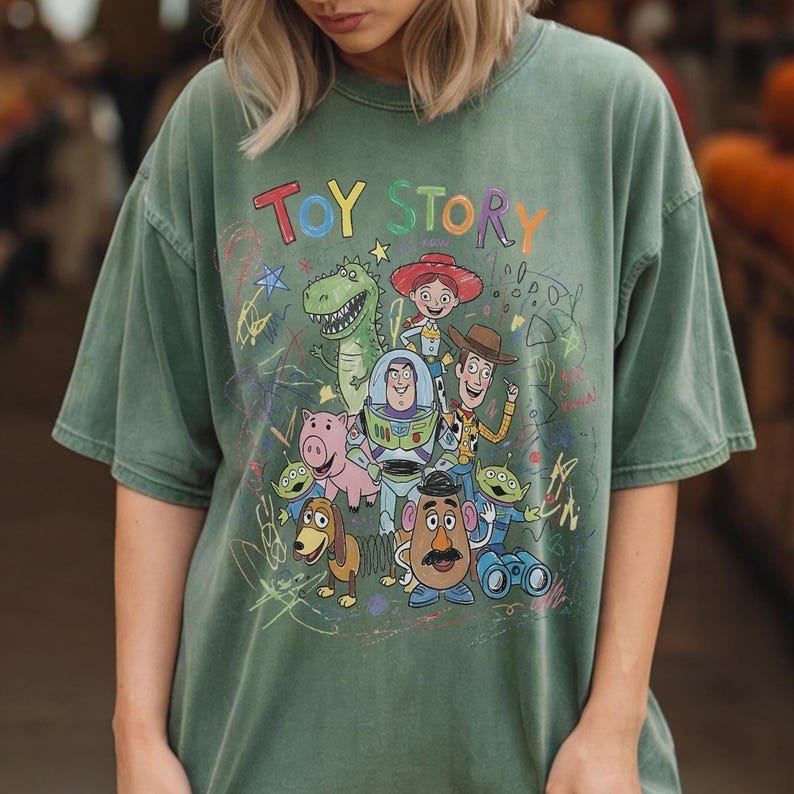 May include: A sage green short-sleeve t-shirt featuring a colourful graphic print of Toy Story characters including Woody, Buzz Lightyear, Jessie, Rex, Hamm, Slinky Dog, Mr. Potato Head, and Aliens. The text TOY STORY appears above the characters.