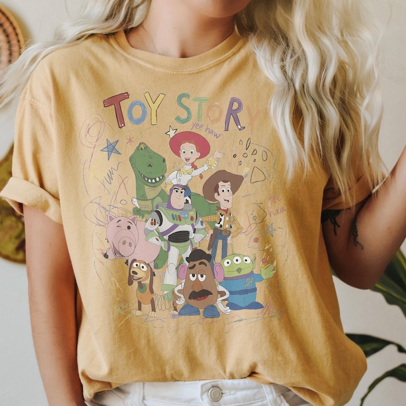 Puede incluir: Camiseta de color amarillo mostaza con un gr&aacute;fico de los personajes de Toy Story, incluyendo a Woody, Buzz Lightyear y Rex. Las palabras "TOY STOR" est&aacute;n impresas en la parte superior en letras coloridas. El dise&ntilde;o tiene una est&eacute;tica vintage y esbozada.