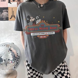 May include: Dark grey t-shirt with a graphic of Mickey Mouse holding a film reel, and the text "The Great Movie Ride" in a marquee style. The shirt also features the text "A Spectacular Journey Into The Movies" and the years "1989-2017".