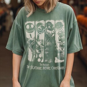 May include: A sage green short-sleeve t-shirt featuring a white graphic print of characters from Tim Burton's The Nightmare Before Christmas. The design includes four vertical panels with character illustrations and the movie title text below.