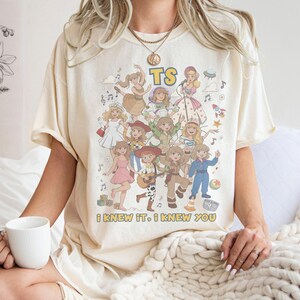 May include: A cream-colored short-sleeve t-shirt featuring a graphic print of multiple cartoon characters in various outfits. The text TS is at the top, and the text I KNEW IT, I KNEW YOU is at the bottom. The design includes musical notes and stars.