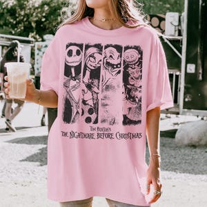 May include: A light pink short-sleeve t-shirt featuring a black graphic print of characters from Tim Burton's The Nightmare Before Christmas. The design includes vertical panels showing Jack Skellington, Sally, Oogie Boogie, and other figures.