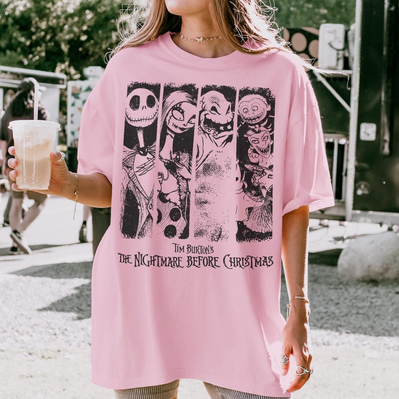 May include: A light pink short-sleeve t-shirt featuring a black graphic print of characters from Tim Burton's The Nightmare Before Christmas. The design includes vertical panels showing Jack Skellington, Sally, Oogie Boogie, and other figures.