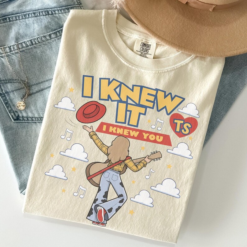 K&ouml;nnte beinhalten: Ein cremefarbenes T-Shirt mit einer Grafik einer langhaarigen Person, die eine Akustikgitarre spielt und ein gelbes Karohemd sowie eine Schlaghose mit Kuhfleckenmuster tr&auml;gt. Der Text lautet I KNEW IT und I KNEW YOU, erg&auml;nzt durch Wolken, Noten, einen roten Hut und ein rotes Herz mit TS.