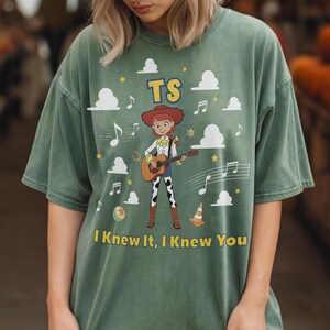 Puede incluir: Una camiseta de manga corta color verde salvia con un gr&aacute;fico de una vaquera tocando una guitarra ac&uacute;stica. El dise&ntilde;o incluye nubes blancas, notas musicales, estrellas, una pelota y un cono. El texto dice TS y I Knew It, I Knew You.