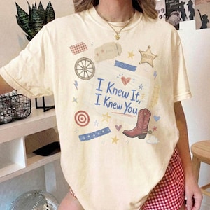 May include: A cream-colored short-sleeve t-shirt featuring a central graphic of a lined notebook page with the blue text I Knew It, I Knew You. The design includes a brown cowboy boot, a wagon wheel, a sheriff star, and various small icons.