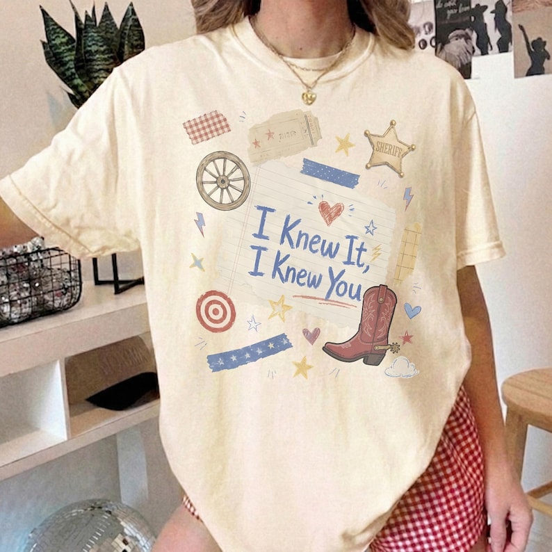 May include: A cream-colored short-sleeve t-shirt featuring a central graphic of a lined notebook page with the blue text I Knew It, I Knew You. The design includes a brown cowboy boot, a wagon wheel, a sheriff star, and various small icons.