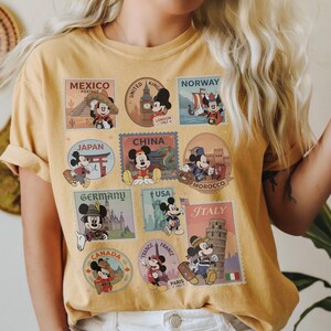 May include: Mustard yellow t-shirt featuring Mickey Mouse in various travel-themed postage stamp designs. Stamps include destinations like Mexico, London, Norway, Japan, China, Morocco, Germany, USA, Italy, Canada, and Paris.