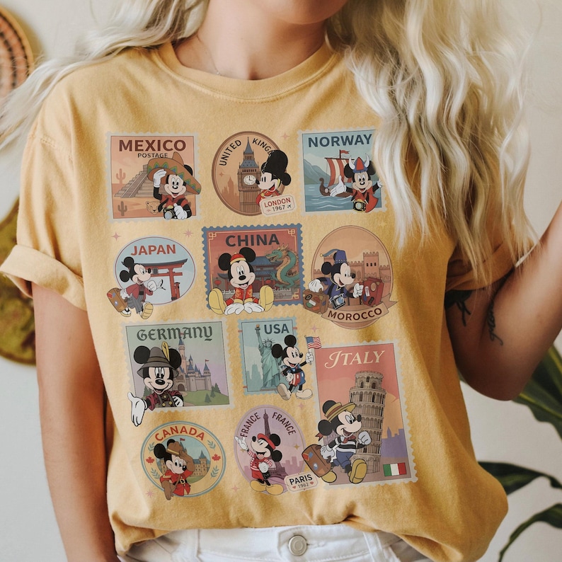 May include: Mustard yellow t-shirt featuring Mickey Mouse in various travel-themed postage stamp designs. Stamps include destinations like Mexico, London, Norway, Japan, China, Morocco, Germany, USA, Italy, Canada, and Paris.
