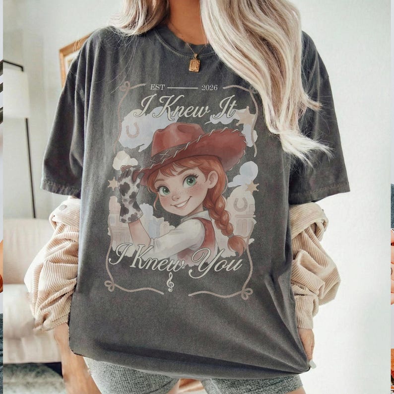 May include: A charcoal gray short-sleeve t-shirt featuring a graphic of a cartoon cowgirl with a red hat and braided hair. The text reads "EST 2026", "I Knew It", and "I Knew You" with decorative western-style line art and horseshoe icons.