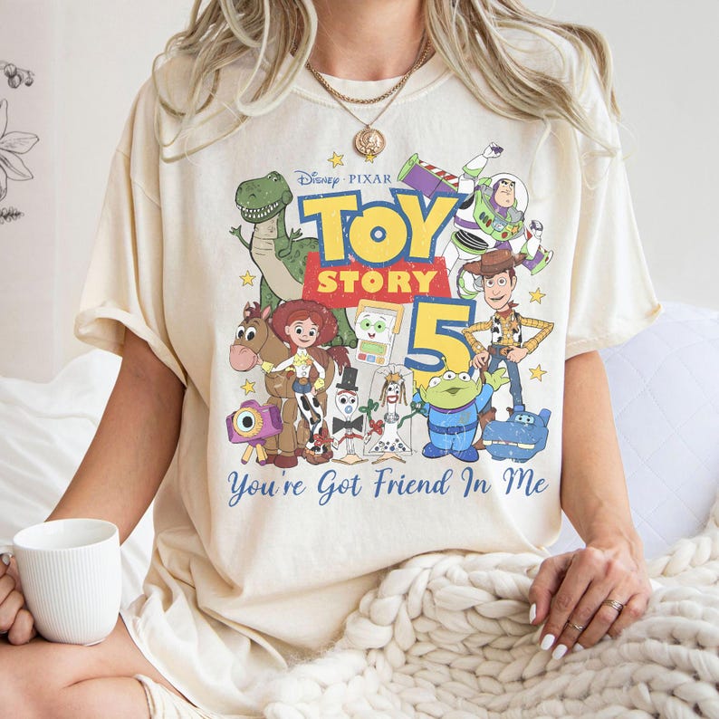 May include: A cream-colored short-sleeve t-shirt featuring a graphic print of Disney Pixar Toy Story characters including Woody, Buzz Lightyear, Jessie, Rex, and others. The text reads Toy Story 5 and You've Got Friend In Me in blue and red.