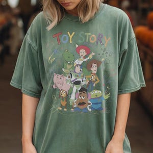 Puede incluir: Camiseta verde con la inscripci&oacute;n "TOY STORY" en letras coloridas y un gr&aacute;fico de los personajes de Toy Story. Los personajes incluyen a Woody, Buzz Lightyear y Rex. La camiseta tiene un aspecto vintage y desgastado.