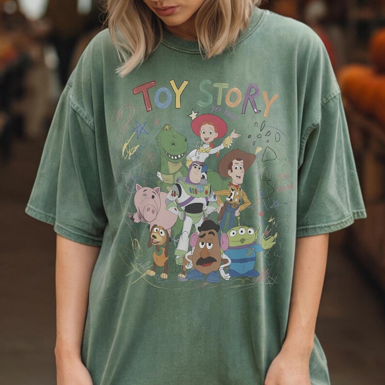 Puede incluir: Camiseta verde con la inscripci&oacute;n "TOY STORY" en letras coloridas y un gr&aacute;fico de los personajes de Toy Story. Los personajes incluyen a Woody, Buzz Lightyear y Rex. La camiseta tiene un aspecto vintage y desgastado.