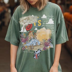 May include: A sage green short-sleeve t-shirt featuring a graphic print of a blonde character in a blue dress and cowboy boots, surrounded by flowers, clouds, and a red hat. The design includes the text "IS" and cursive script lyrics.