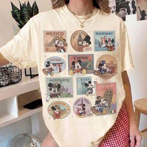 May include: Cream-colored t-shirt featuring Mickey Mouse postage stamp designs from various countries, including Mexico, Japan, China, and Italy. Each stamp showcases Mickey in different outfits and settings, with country names and landmarks.
