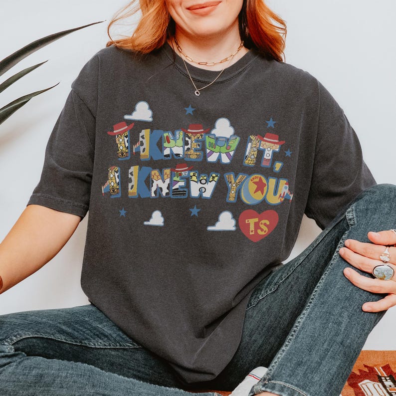 K&ouml;nnte beinhalten: Ein anthrazitfarbenes Kurzarm-T-Shirt mit dem Text I KNEW IT, I KNEW YOU in einer mehrfarbigen Schriftart mit charakterbezogenen Mustern. Das Design enth&auml;lt kleine wei&szlig;e Wolken, blaue Sterne und ein rotes Herz mit den Buchstaben TS darin.