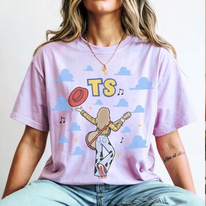 May include: A light purple short-sleeve t-shirt featuring a graphic of a person with long hair wearing a yellow plaid shirt and cow-print pants, holding a guitar and a red hat. The design includes blue clouds, yellow stars, music notes, and the text TS.