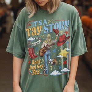 K&ouml;nnte beinhalten: Ein salbeigr&uuml;nes Kurzarm-T-Shirt mit einem Grafikdruck mit dem Text IT'S A TAY STORY und Baby Just Say Yes. Das Design zeigt eine Person mit Gitarre, rote Cowboystiefel, rote H&uuml;te, ein Perlenarmband, Sterne und Wolken auf dem Stoff.