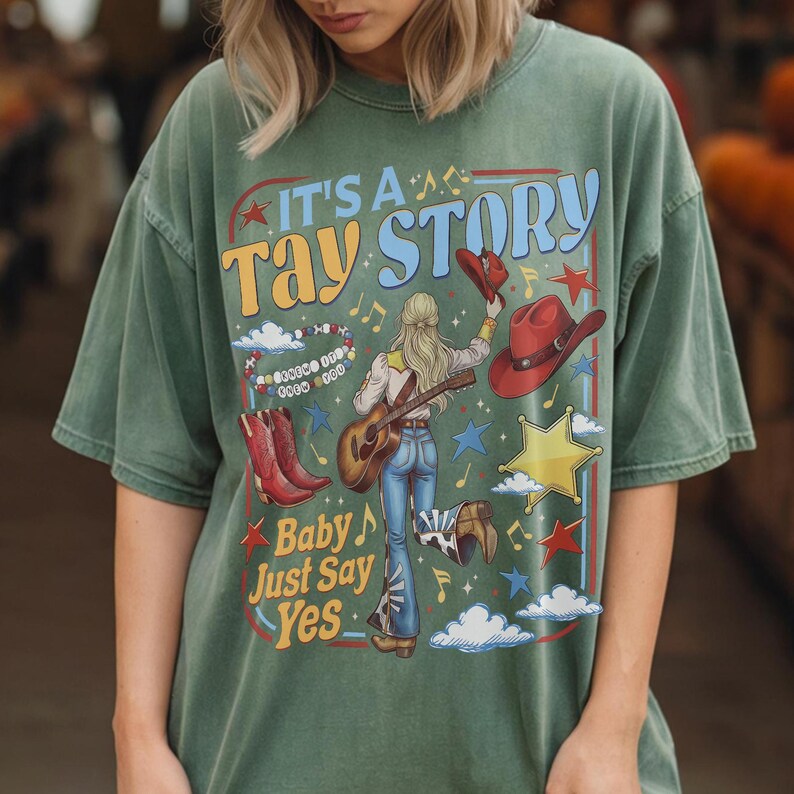 K&ouml;nnte beinhalten: Ein salbeigr&uuml;nes Kurzarm-T-Shirt mit einem Grafikdruck mit dem Text IT'S A TAY STORY und Baby Just Say Yes. Das Design zeigt eine Person mit Gitarre, rote Cowboystiefel, rote H&uuml;te, ein Perlenarmband, Sterne und Wolken auf dem Stoff.