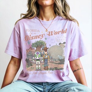May include: A light purple t-shirt featuring a Disney World graphic. The graphic includes the text "Florida Est. 1971 Disney World" and images of Buzz Lightyear, Woody, and various park landmarks. The shirt also has the text "The Most Magical Place On Earth."