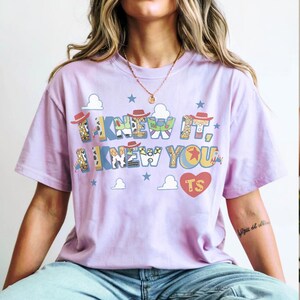 K&ouml;nnte beinhalten: Ein hellviolettes Kurzarm-T-Shirt mit dem Text I KNEW IT, I KNEW YOU in einer mehrfarbigen Musterschrift. Das Design enth&auml;lt kleine Cowboyh&uuml;te, blaue Wolken, Sterne und ein rotes Herz mit den gelb gedruckten Buchstaben TS.