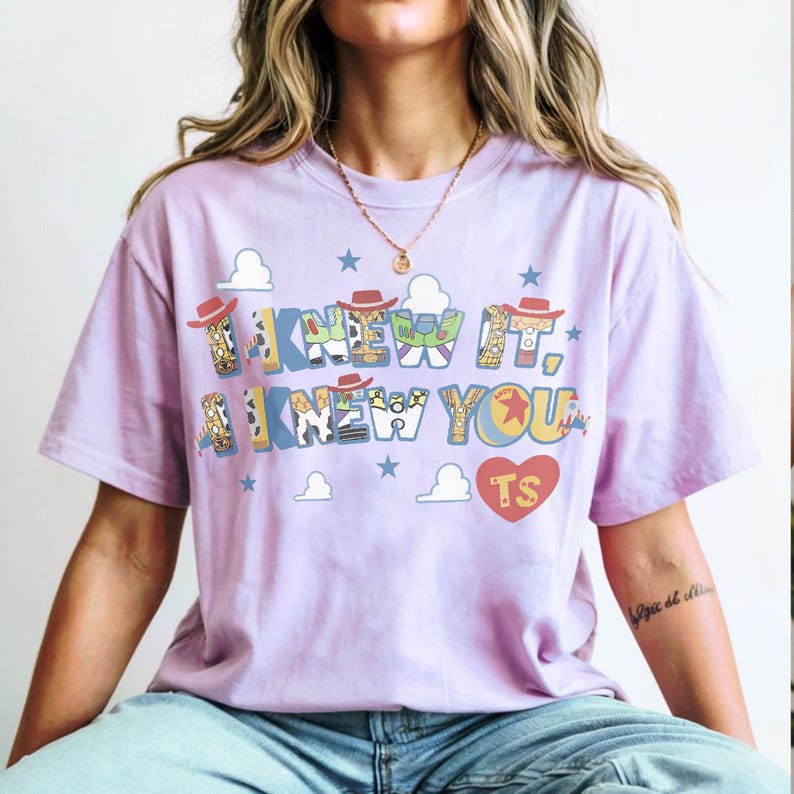 K&ouml;nnte beinhalten: Ein hellviolettes Kurzarm-T-Shirt mit dem Text I KNEW IT, I KNEW YOU in einer mehrfarbigen Musterschrift. Das Design enth&auml;lt kleine Cowboyh&uuml;te, blaue Wolken, Sterne und ein rotes Herz mit den gelb gedruckten Buchstaben TS.