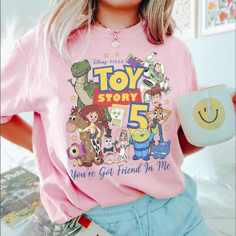 May include: A light pink t-shirt featuring a colorful Disney Pixar Toy Story 5 graphic print. The design includes characters like Rex, Buzz Lightyear, Woody, Jessie, Bullseye, and Forky, with the text Toy Story 5 and You've Got Friend In Me.