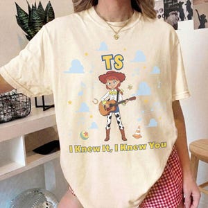 Puede incluir: Una camiseta de manga corta color crema con un gr&aacute;fico de un personaje vaquero tocando una guitarra ac&uacute;stica. El dise&ntilde;o incluye el texto "TS" sobre el personaje y "I Knew It, I Knew You" debajo, rodeado de nubes azules, estrellas y notas musicales.