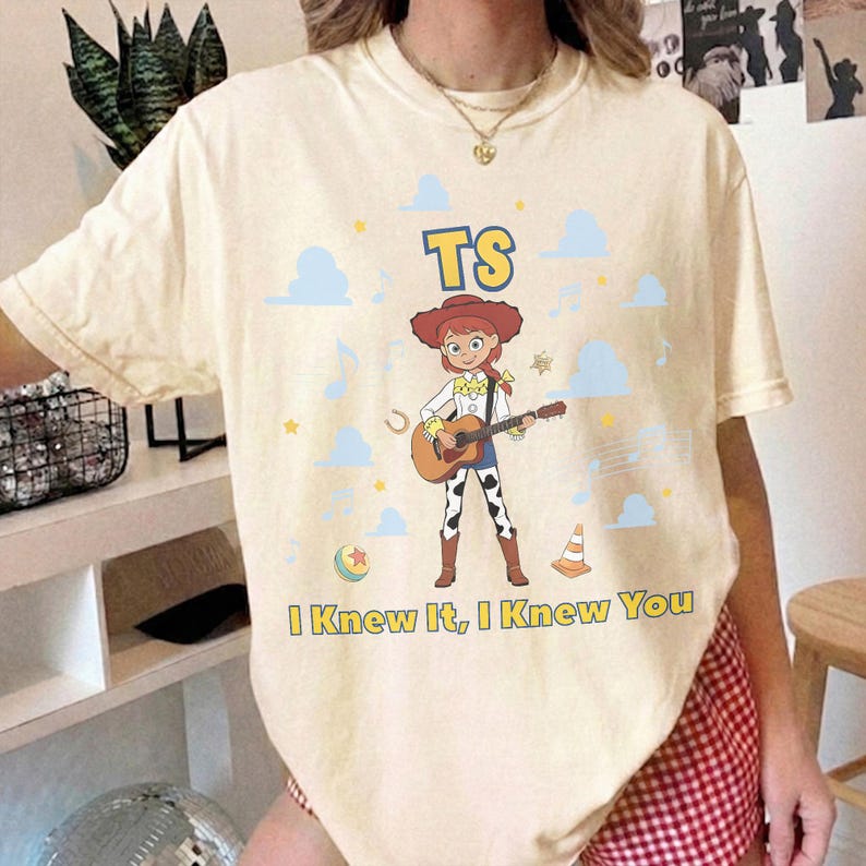 Puede incluir: Una camiseta de manga corta color crema con un gr&aacute;fico de un personaje vaquero tocando una guitarra ac&uacute;stica. El dise&ntilde;o incluye el texto "TS" sobre el personaje y "I Knew It, I Knew You" debajo, rodeado de nubes azules, estrellas y notas musicales.