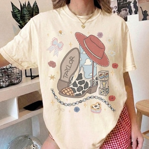 May include: A cream-coloured t-shirt featuring a graphic print of a cowboy boot with a cow print pattern, a red cowboy hat, the name Taylor, the number 13, stacked blocks spelling TS, and a beaded string with the text I KNEW IT YOU KNEW YOU.