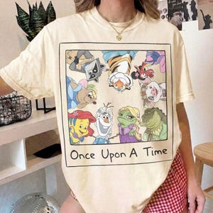 May include: Cream-colored t-shirt featuring a framed graphic of cartoon characters, including a raccoon, tiger, and pig. The phrase "Once Upon A Time" is printed below the graphic in black. The shirt has short sleeves and a crew neck.
