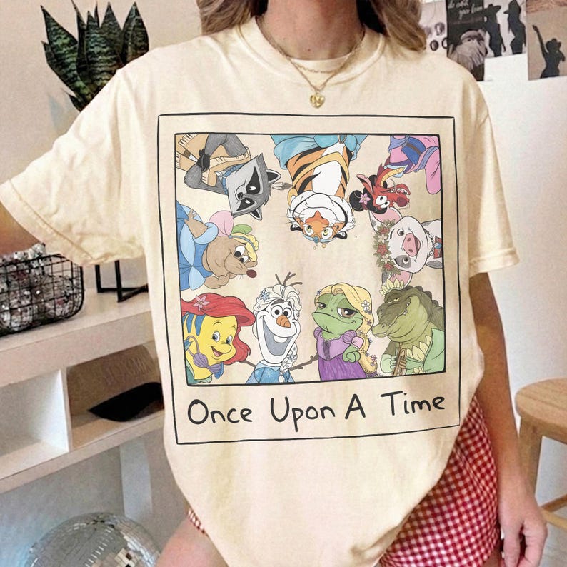 May include: Cream-colored t-shirt featuring a framed graphic of cartoon characters, including a raccoon, tiger, and pig. The phrase "Once Upon A Time" is printed below the graphic in black. The shirt has short sleeves and a crew neck.