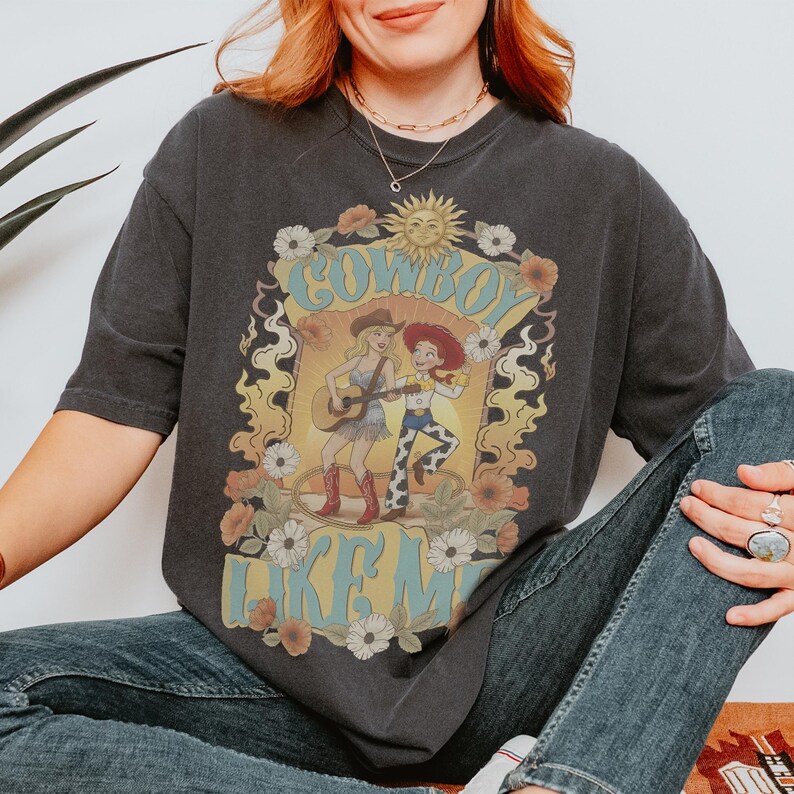 May include: A charcoal gray short-sleeve t-shirt featuring a graphic print of two female characters, one playing an acoustic guitar, surrounded by a floral border, a sun icon, and the text "COWBOY LIKE ME" in a light blue, stylized font.