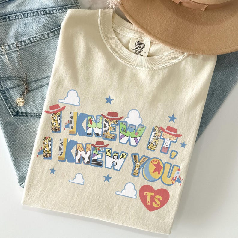 K&ouml;nnte beinhalten: Ein cremefarbenes Kurzarm-T-Shirt mit dem Text I KNEW IT, I KNEW YOU in blauen Blockbuchstaben, die mit Mustern von Cowboy- und Weltraum-Ranger-Figuren gef&uuml;llt sind. Das Design enth&auml;lt kleine blaue Sterne, wei&szlig;e Wolken und ein rotes Herz mit TS.