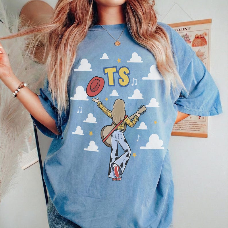 May include: A light blue t-shirt featuring a graphic of a person with long hair wearing a yellow plaid shirt and cow-print pants, holding a guitar. The design includes a red hat, white clouds, yellow stars, musical notes, and the text TS in yellow.