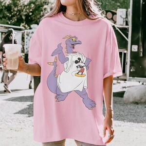 Peut inclure: T-shirt rose clair ample avec un dragon violet portant un t-shirt blanc avec un motif Mickey Mouse. Le dragon a des cornes jaunes et une longue queue. Le t-shirt est de style d&eacute;contract&eacute; et confortable.