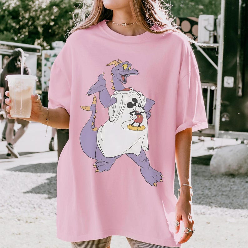 Peut inclure: T-shirt rose clair ample avec un dragon violet portant un t-shirt blanc avec un motif Mickey Mouse. Le dragon a des cornes jaunes et une longue queue. Le t-shirt est de style d&eacute;contract&eacute; et confortable.
